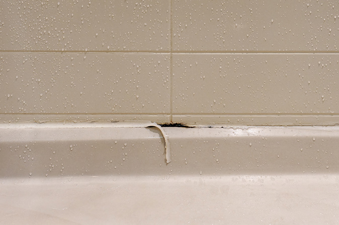 White shower stall with peeling caulk and mold growing underneath it.