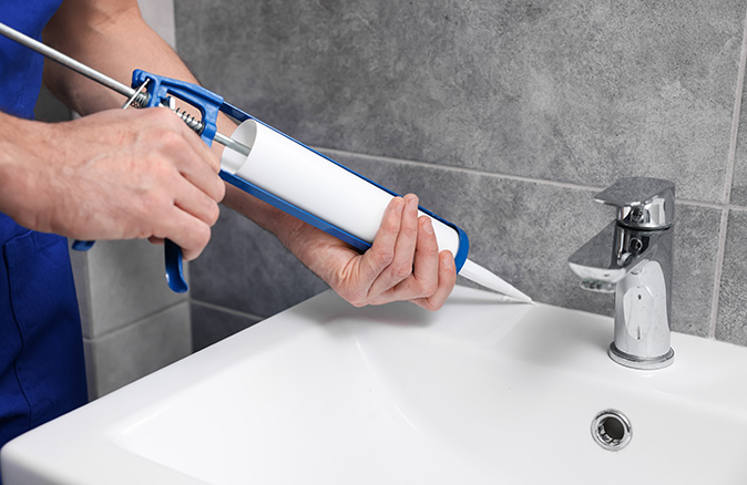 A close-up of a man using a caulking gun to seal a sink in a bathroom.